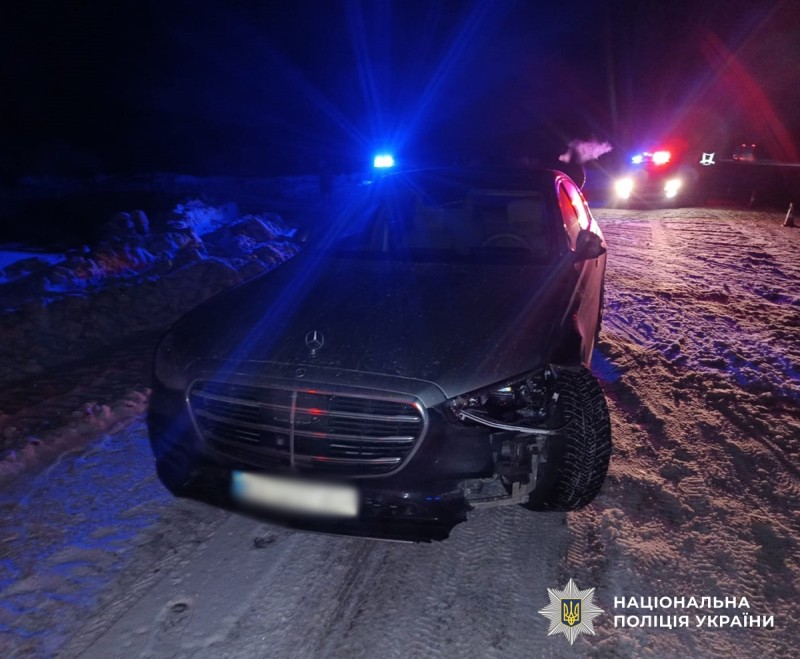 Місце події дорожньо-транспортної пригоди. Легковий автомобіль темного кольору, стоїть на засніженій дорозі в темну пору доби. Передня частина машини пошкоджена. Позаду автомобіля з увімкненими проблисковими маячками синього й червоного кольорів, стоять два службові авто поліцейських.