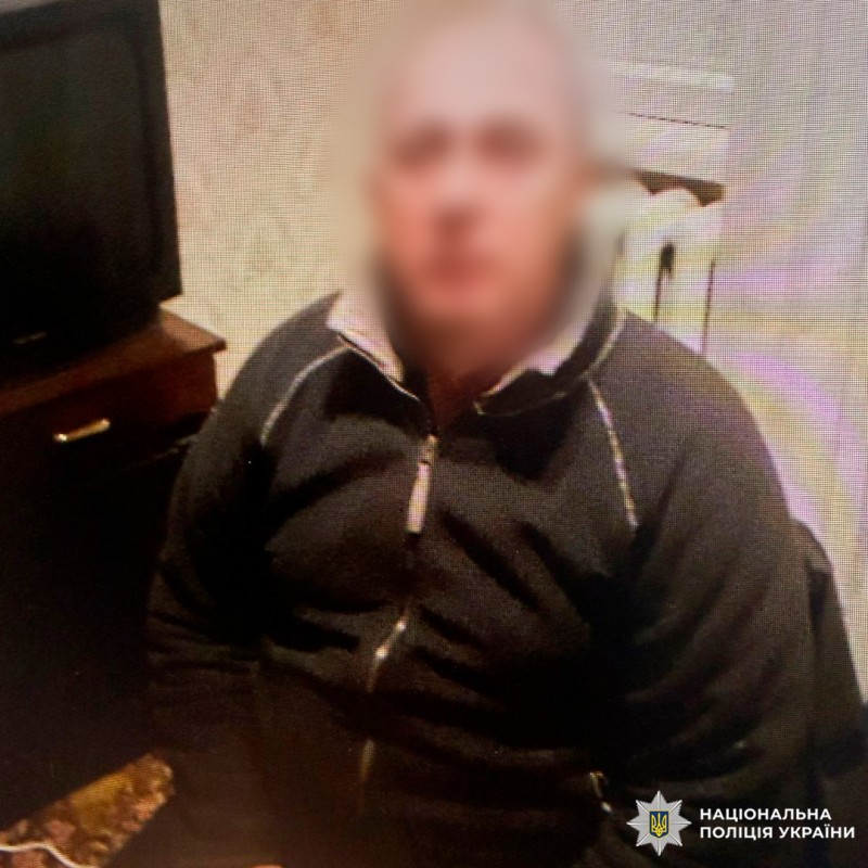 Чоловік у темній куртці сидить у кімнаті з телевізором на шафі, батареєю та шпалерами з візерунком.