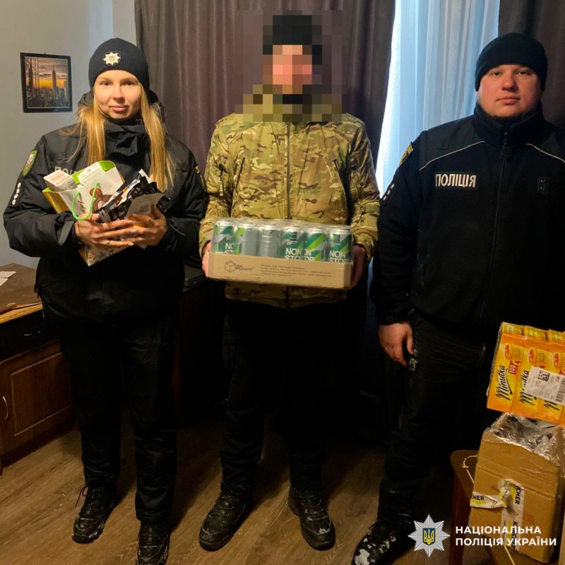 У приміщенні троє осіб: двоє поліцейських у чорній формі з написом «Поліція» та один чоловік у камуфляжній куртці. Він тримає коробку з банками «NON STOP». Поліцейський ліворуч тримає паковані продукти, праворуч - коробка з напоями «Mirinda».