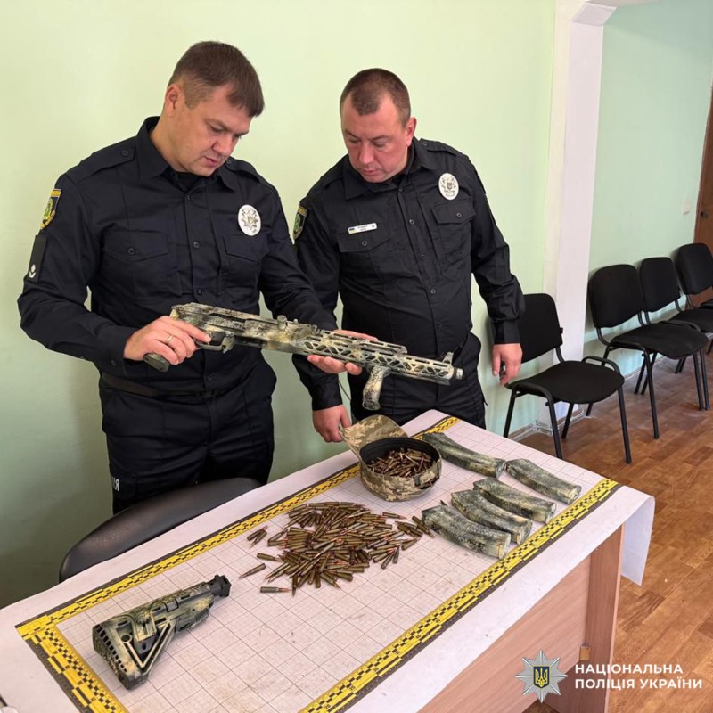Двоє поліцейських у формі Національної поліції України оглядають зброю та боєприпаси, розкладені на столі. Серед предметів камуфльований автомат, магазини та велика кількість набоїв.