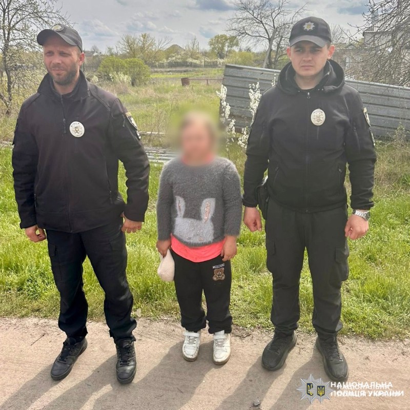 Двоє поліцейських у чорній формі стоять на вулиці поруч із дитиною у сірому светрі з малюнком.