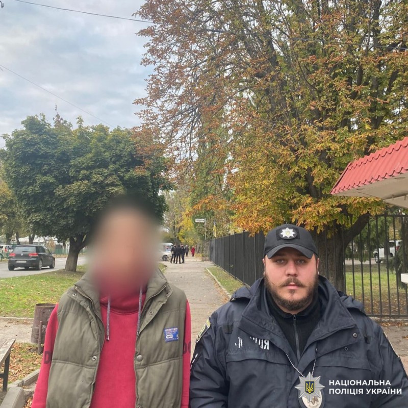 Поліцейський з затриманним який одягнений в червону худі та зелену жилетку, на тротуарі серед осінніх дерев.
