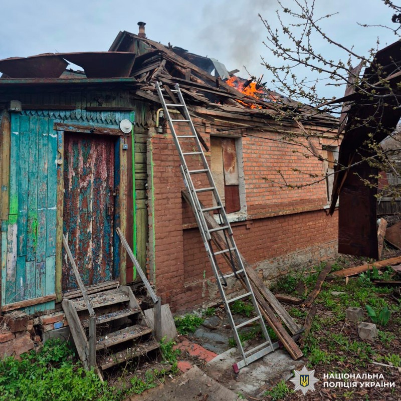 Пошкоджений житловий будинок із частково зруйнованим дахом, охопленим вогнем та димом; на фасаді встановлена металева драбина, територія засмічена уламками.