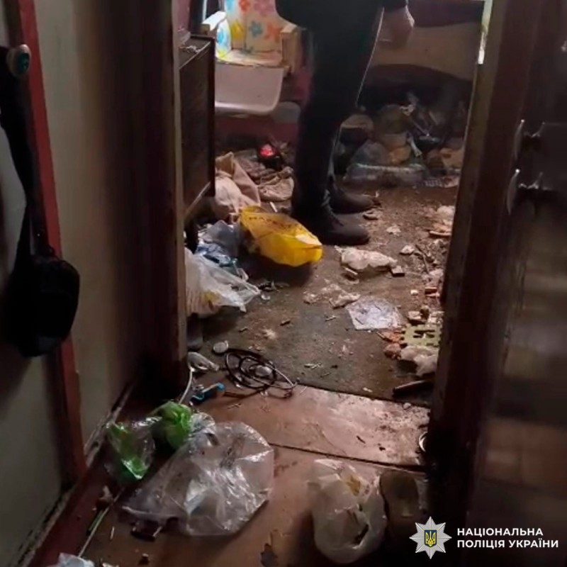 Зображено внутрішнє приміщення з ознаками занедбаності: підлога вкрита побутовими відходами, у дверному проході стоїть особа в темному одязі. Стіни та підлога мають видимі пошкодження. За дверима подальший безлад і зламані предмети.