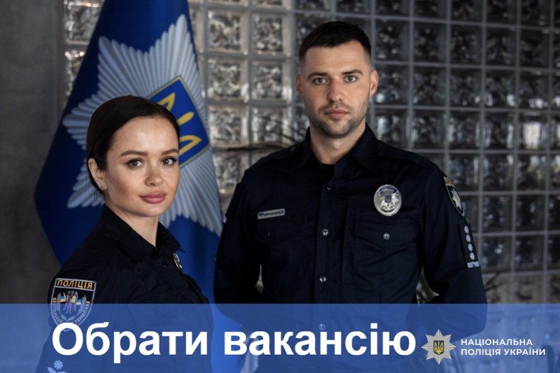 Двоє поліцейських у формі стоять перед стягом НПУ на фоні скляної стіни; внизу — банер із написом "Обрати вакансію", що вказує на рекрутингову кампанію.