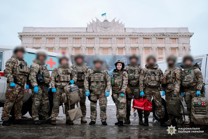 Група співробітників у камуфляжній формі з медичними сумками та спорядженням стоїть перед урядовою будівлею. На задньому плані - автомобілі швидкої допомоги.
