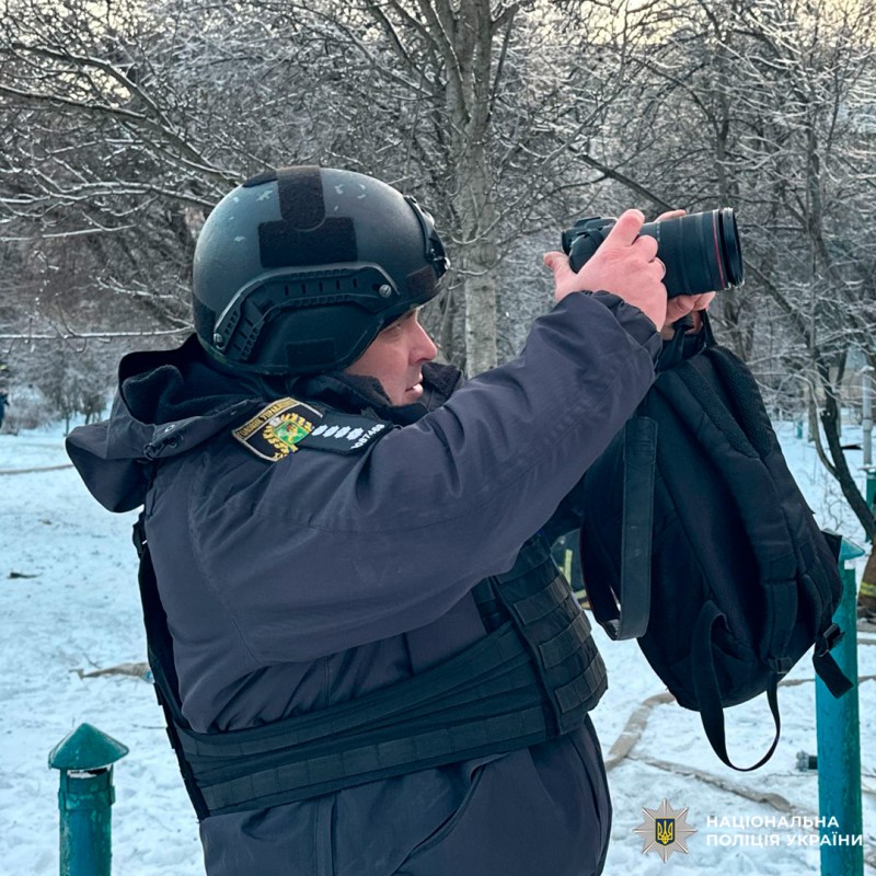 Співробітник Національної поліції України у зимовому спорядженні фотографує місце події в засніженому парку.