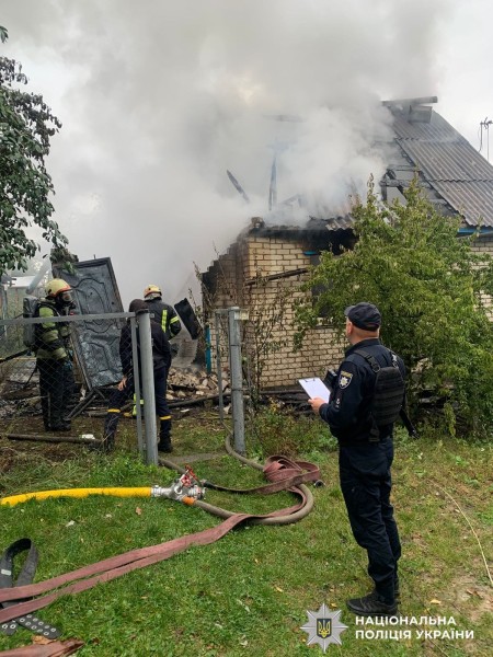 На фото зображений поліцейський та працівники ДСНС, які працюють на місці події де триває пожежеа у будинку.