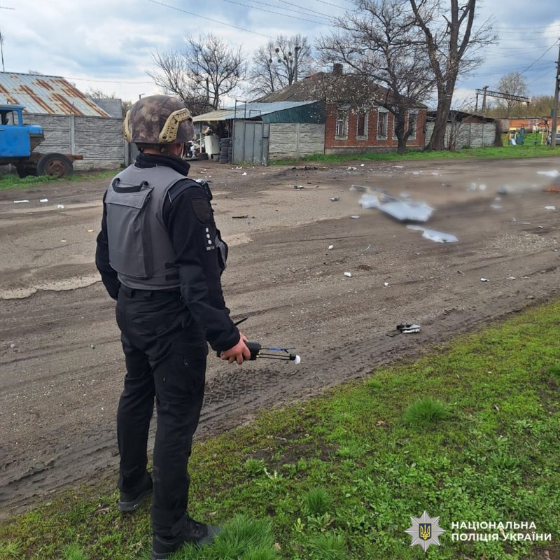 Поліцейський у захисному спорядженні проводить огляд пошкодженої території з уламками, на тлі будівель та службового транспорту.