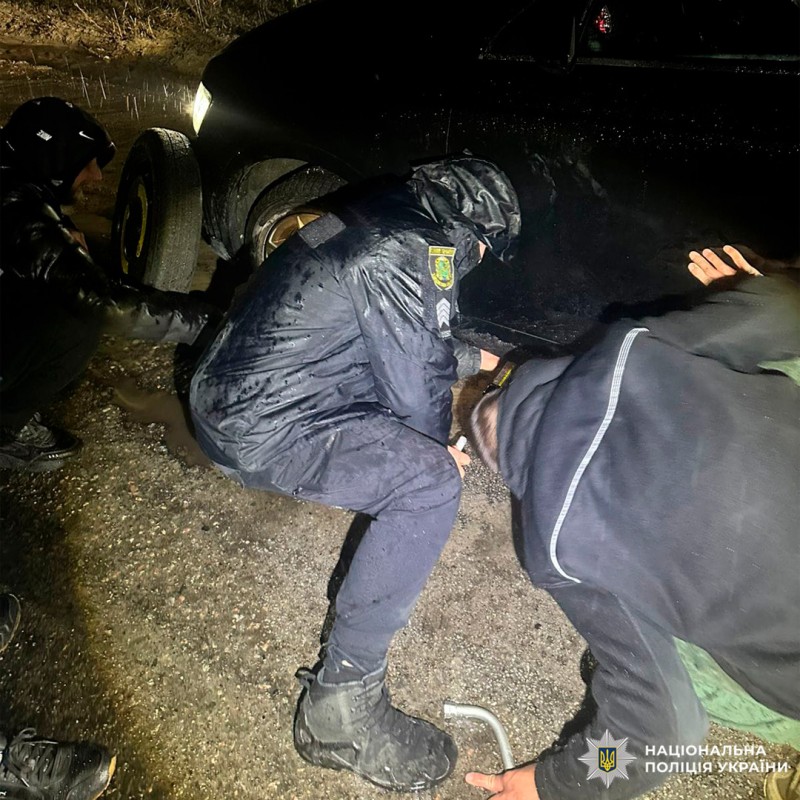 Поліцейські та громадяни змінюють колесо автомобіля під час дощу вночі.