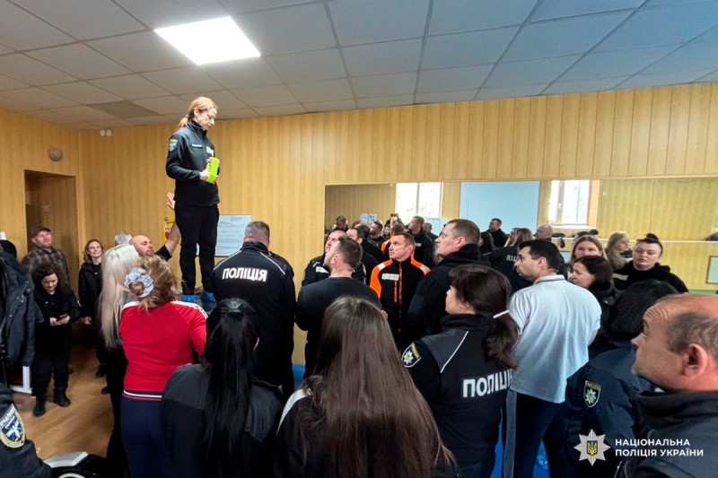 На фото зображено навчальний або командний захід Національної поліції України. У приміщенні з дерев’яними панелями на стінах стоїть група поліцейських у формі з написом «ПОЛІЦІЯ». Один із учасників у спортивному одязі — стоїть на підвищенні, тримаючи зелений предмет у руках, і звертається до присутніх.