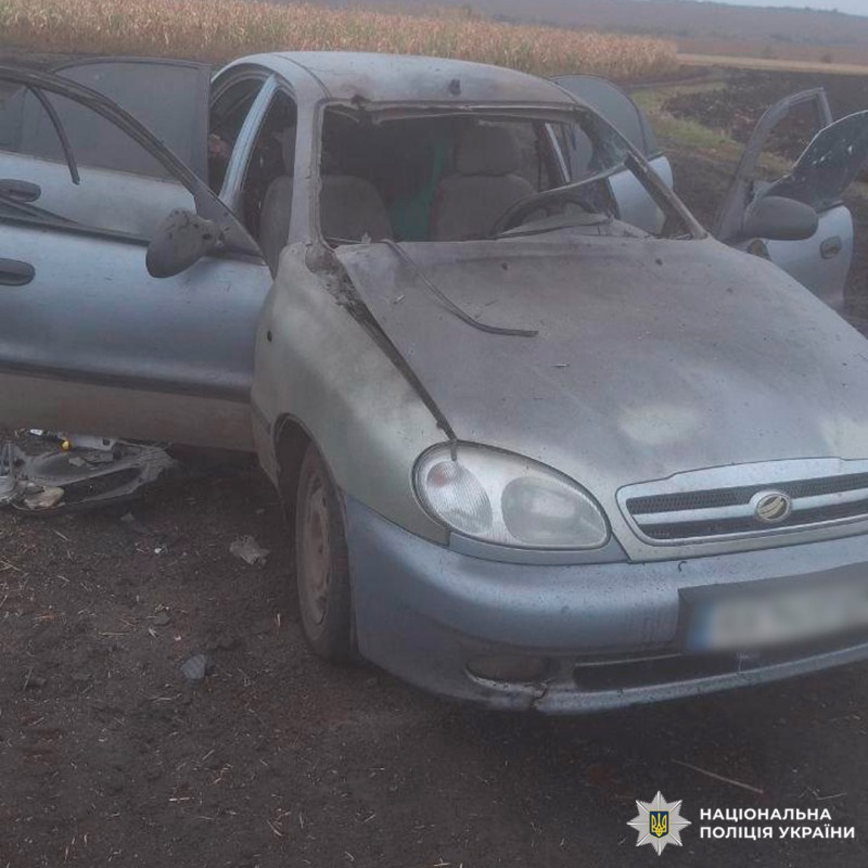 Пошкоджений сріблястий седан з українськими номерами стоїть на польовій дорозі. Розбите лобове скло, вм'ятий дах і відкрита дверцята.