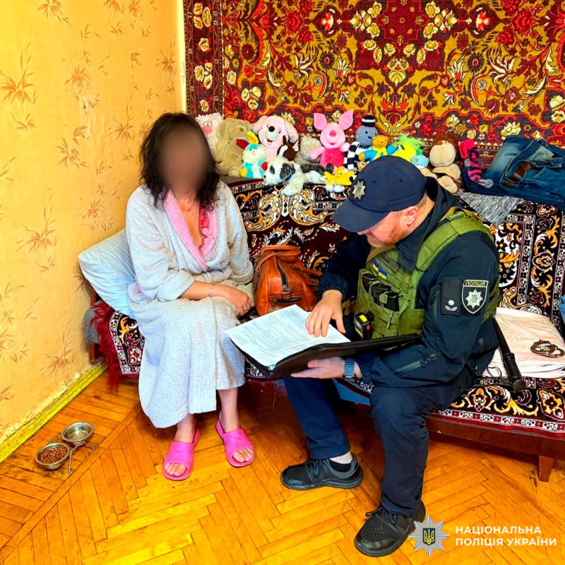 Жінка в халаті та рожевих капцях сидить на дивані в кімнаті з дерев’яною підлогою та червоним килимом на стіні. Поруч із нею співробітник Національної поліції України у формі, який заповнює документи. На дивані  м’які іграшки та особисті речі, на підлозі миски для домашніх тварин.