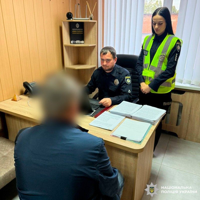 двоє поліцейських, один у формі за ноутбуком, інша у жилеті ПОЛІЦІЯ, стоїть поруч; цивільна особа сидить навпроти, ймовірно, проходить допит. На столі документи, телефон.