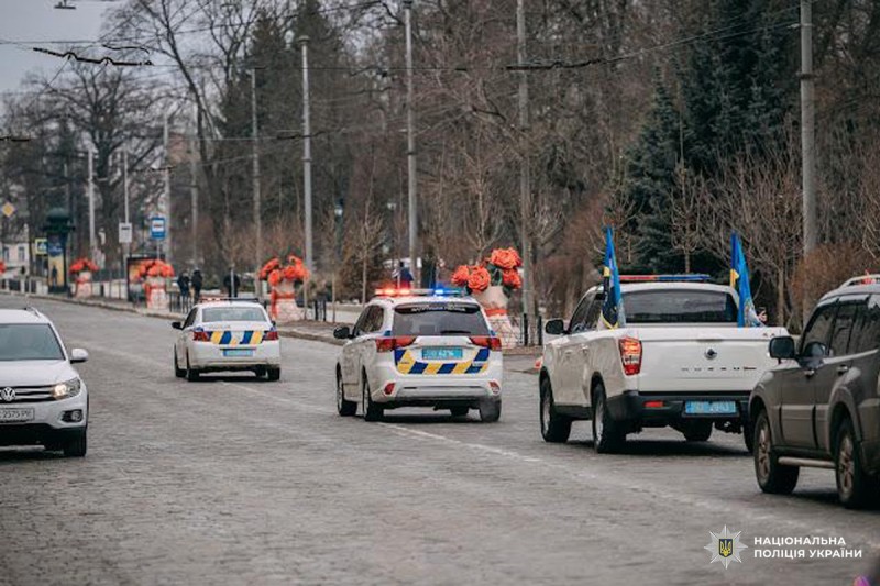 Колона службових автомобілів Національної поліції України з увімкненими проблисковими маячками рухається центральною вулицею міста. Попереду  пікап із державними прапорами.