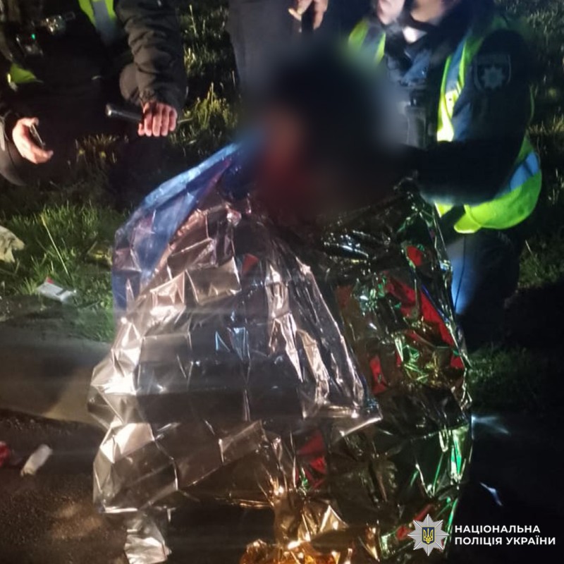 Рятувальники надають допомогу постраждалому, загорнутому в термоковдру, на узбіччі дороги. Людина сидить на землі, отримуючи медичну підтримку. Працівники одягнені у світловідбивальні жилети.