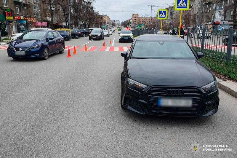На передньому плані — чорний Audi, ліворуч — синій Nissan. Біля пішохідного переходу з червоно-білою розміткою встановлені конуси, поруч — поліцейський автомобіль із маячками. На задньому плані — будівлі та пішоходи.