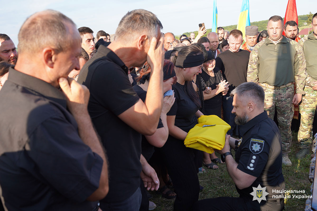 Дружині загиблого поліцейського під час поховання керівник підрозділу поліції вручає державний прапор України, стоячи на колінах