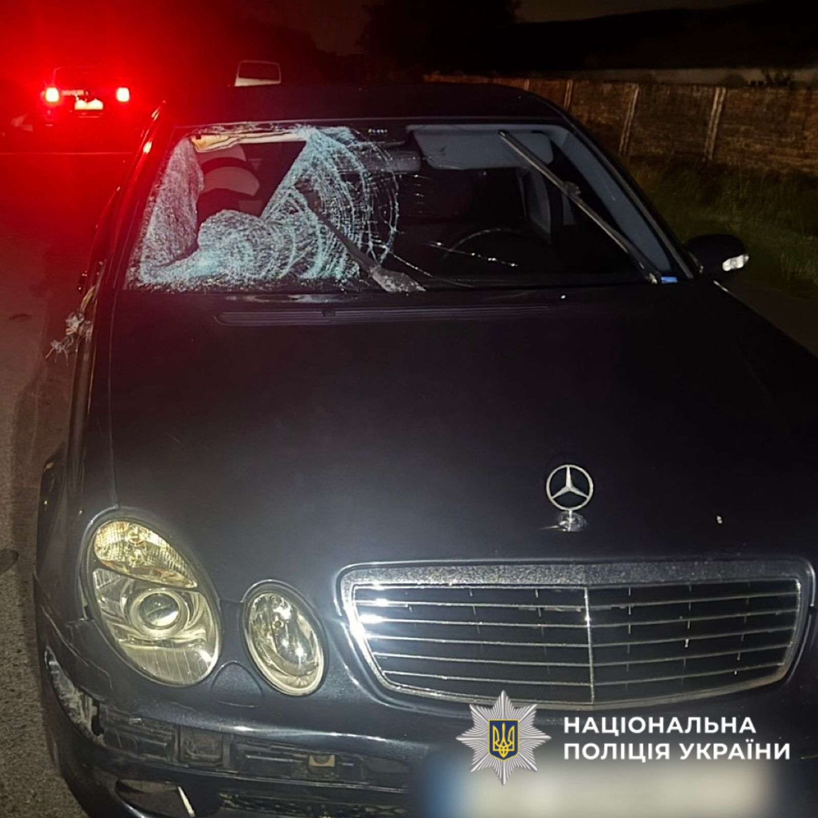 Чорний легковий автомобіль "Mercedes-Benz" із сильно пошкодженим лобовим склом — у ньому велика тріщина від удару. Машина стоїть на дорозі в темну пору доби. У нижньому правому куті зображений логотип Національної поліції України.