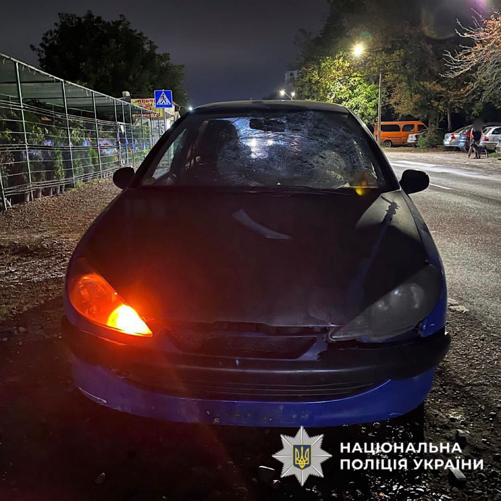 Автомобіль із увімкненою лівою фарою стоїть на узбіччі дороги в темну пору доби. На лобовому склі значні тріщини від удару. У нижньому правому куті зображено логотип Національної поліції України.