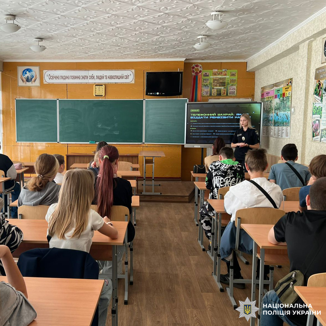 Класна кімната з учнями за партами, викладач у формі поліції проводить урок перед дошкою та екраном.