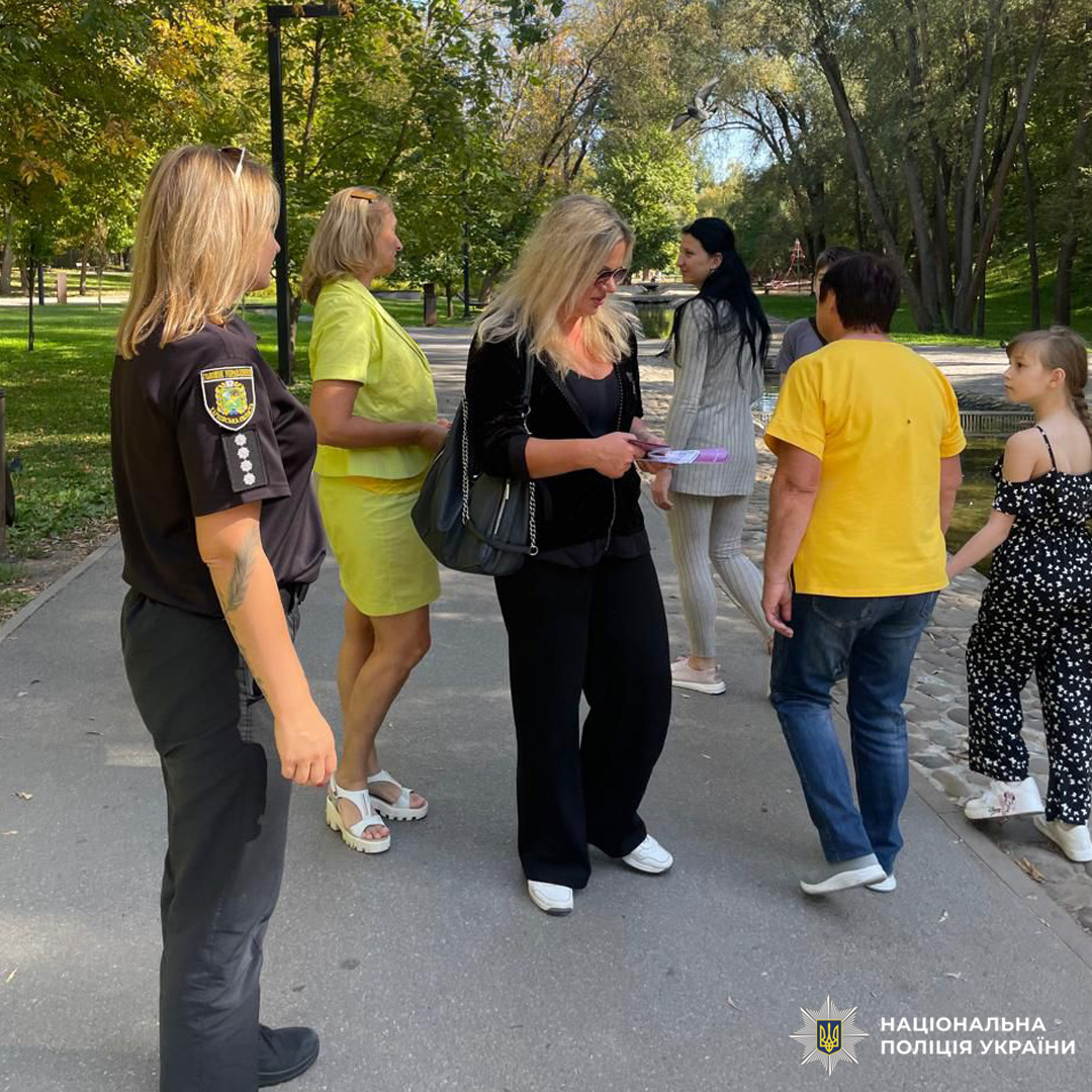 Поліцейська спілкується з групою людей у зеленому парку, тримаючи документи.