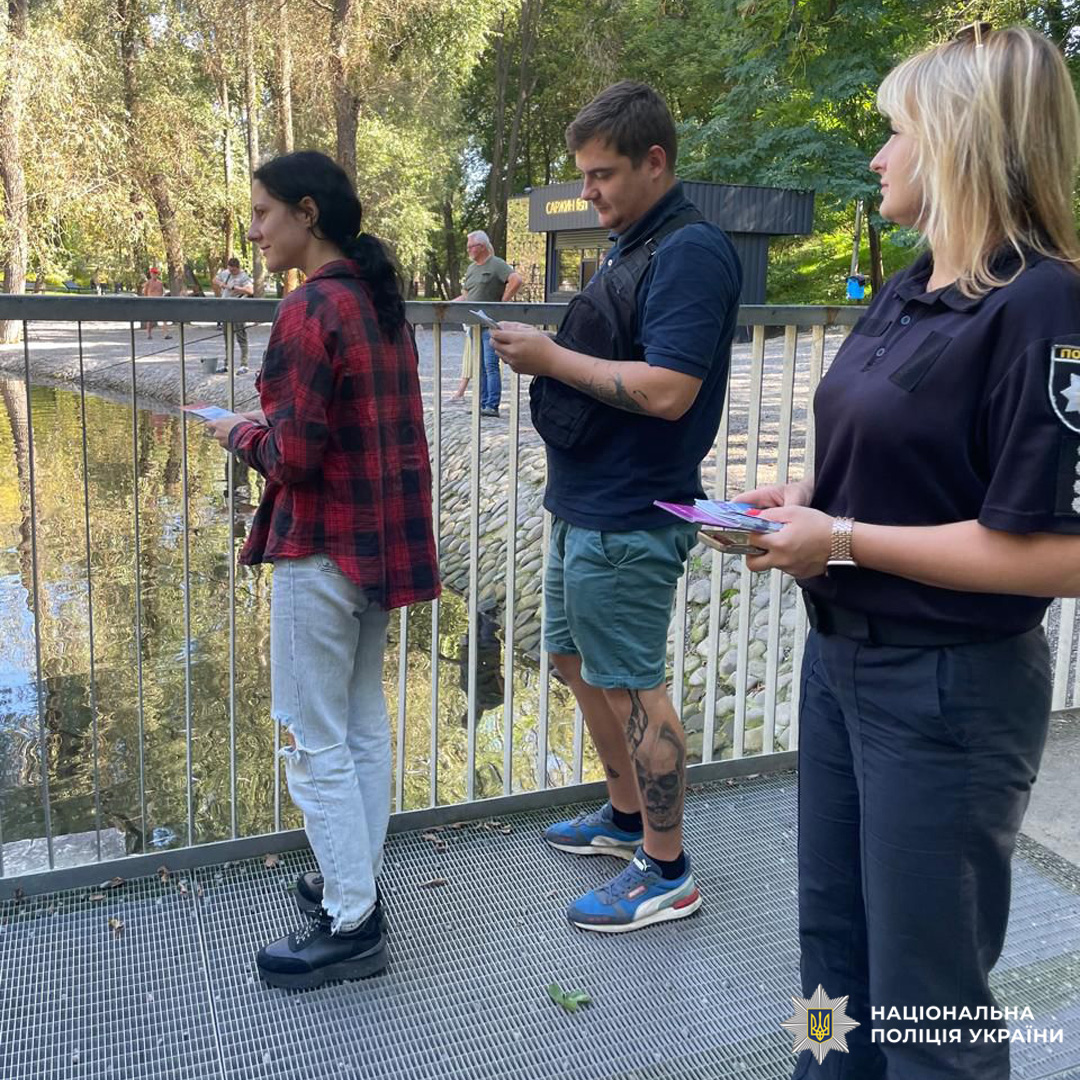 Поліцейська спілкується з двома людьми на мосту над водою в парку, тримаючи документи.