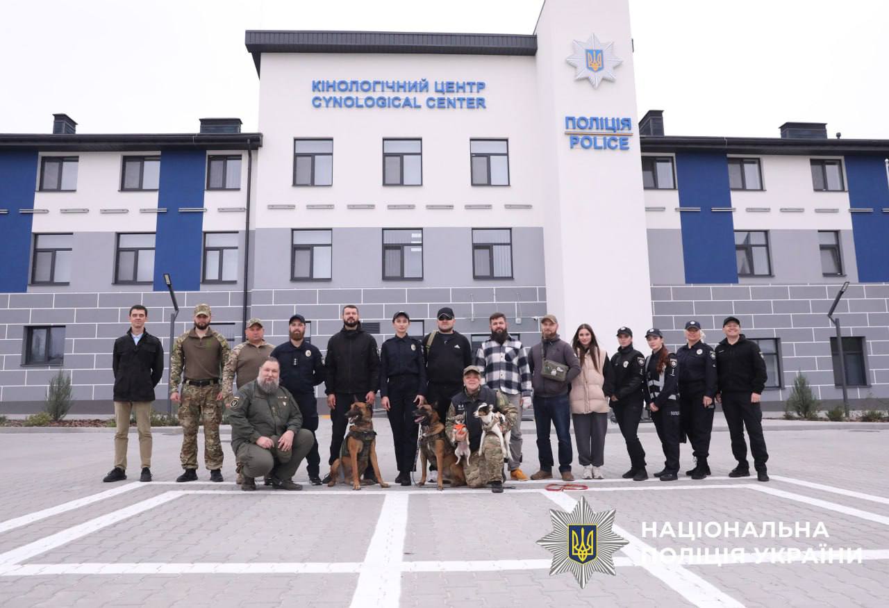 На фото зображені 16 учасників зустрічі, які перебувають у дворі нового Кінологічного центру Національної поліції разом зі службовими собаками.