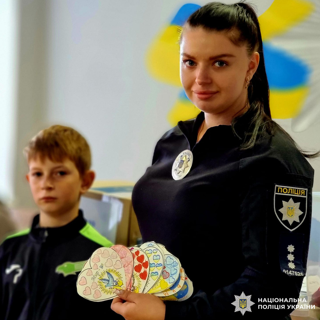 Поліцейська тримає дитячі малюнки у вигляді сердець, на фоні український прапор та хлопчик у спортивному костюмі.