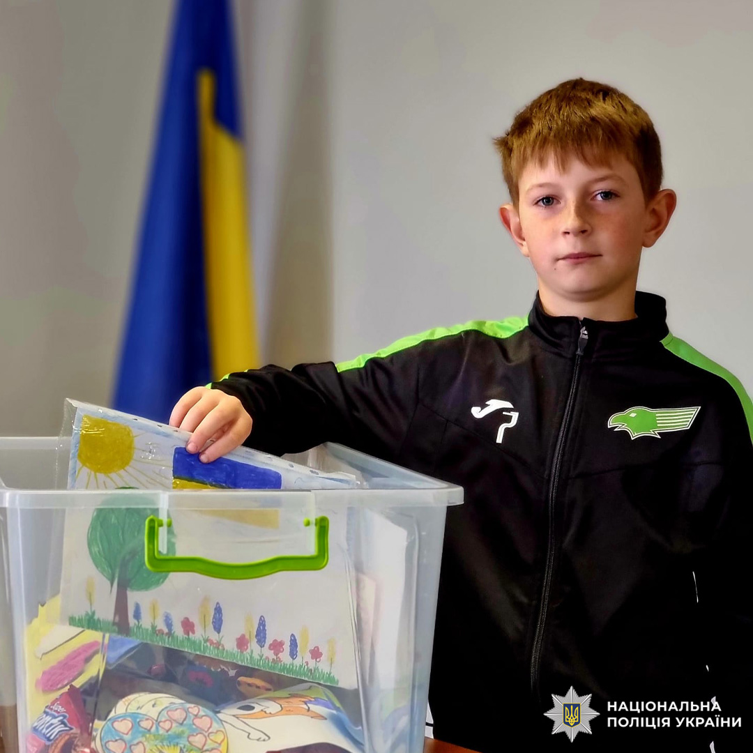 Хлопчик у спортивному костюмі кладе дитячий малюнок у коробку, на фоні український прапор.