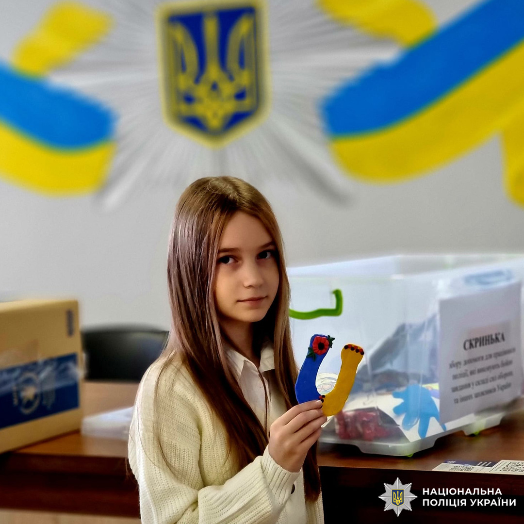 Дівчинка в светрі тримає дитячу виріб у формі літери "U", на фоні емблеми НПУ з українським прапором та скриньки побажань.