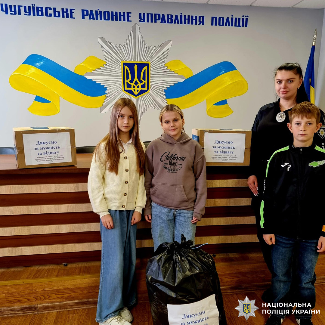 Діти та поліцейська з коробками та пакетом з написами "Дякую за мужність та відвагу" у приміщенні поліції з українською символікою.