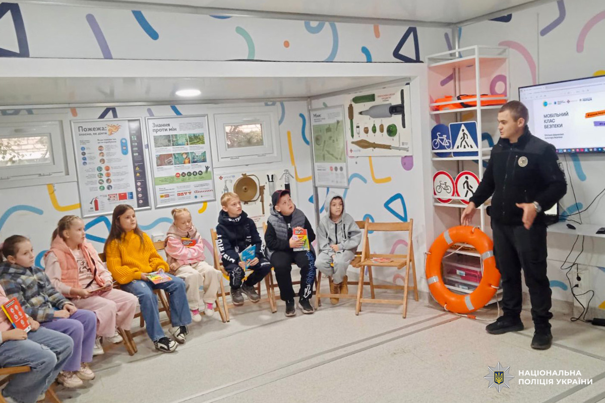 Поліцейський проводить заняття для дітей, які сидять із книгами, у приміщенні з навчальними стендами та рятувальним кругом.