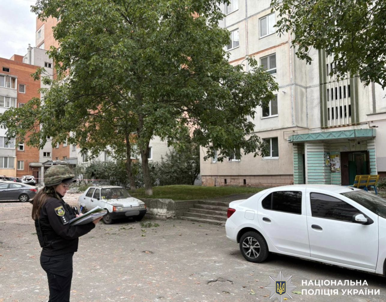 На фото зображена поліцейська, яка стоїть у дворі багатоповерхового будинку та документує наслідки атаки рф.