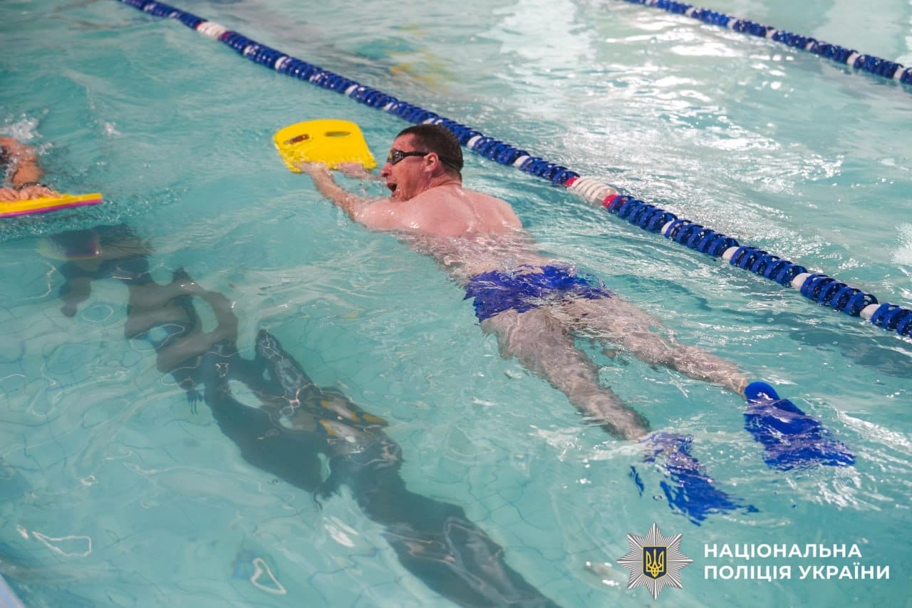 На фотографії зображений чоловік, який пливе у басейні.
Він лежить на воді обличчям вниз і використовує жовту плавальну дошку (кікборд), яку тримає витягнутими руками. На ньому сині плавки та ласти на ногах, а також окуляри для плавання. Чоловік займається плаванням.
Праворуч від нього видно розділову доріжку з синьо-білими поплавками, що вказує на те, що він пливе по своїй доріжці.
