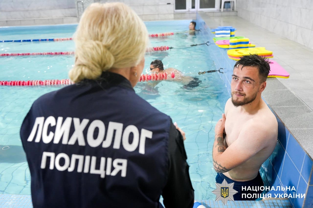 На фотографії зображено спілкування поліцейської-психолога з пораненим захисником. Чоловіка стоїть у басейні, занурений приблизно до грудей, і розмовляє з поліцейською , яка стоїть до нього спиною, поза басейном.
На спині жінки видно напис великими літерами "ПСИХОЛОГ ПОЛІЦІЯ".