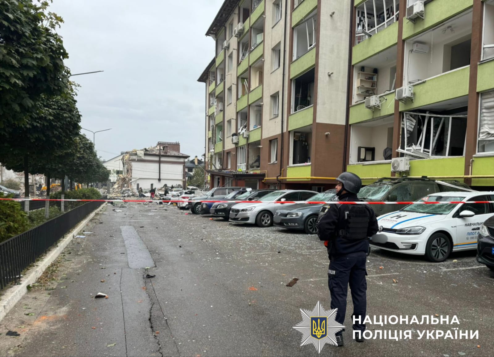 На фото зображений поліцейський, який стоїть на фоні зруйнованого багатоповерхового будинку.