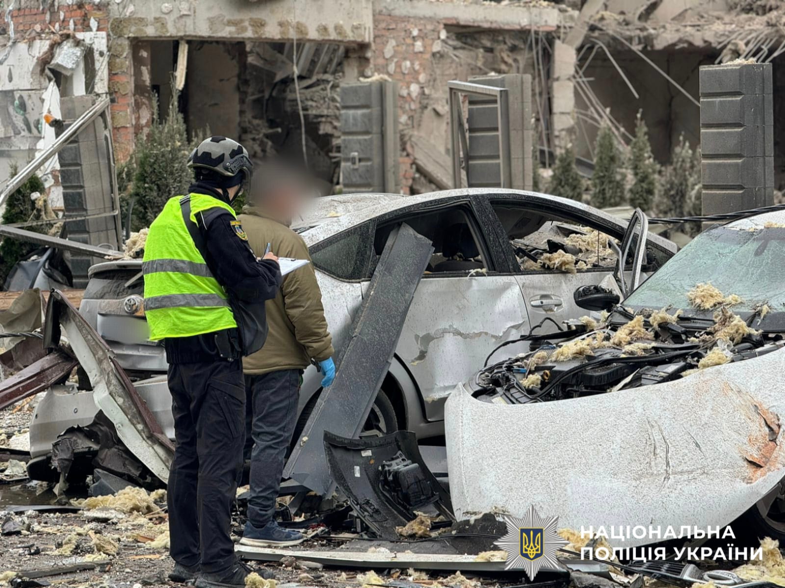 На фото поліцейський та лодин з потерпілих, на фоні зруйнованих автомоілів. Фото з місця події, зроблене під час документування воєнного злочину.