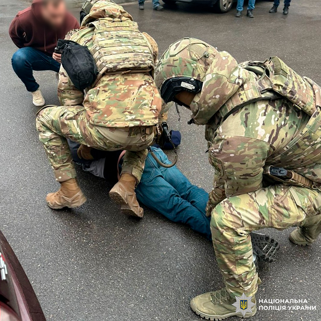 Двоє осіб у військовій або тактичній формі затримують людину, що лежить обличчям донизу на асфальті. Один з них стоїть на спині затриманого, інший тримає його ноги. Сцена відбувається на відкритому майданчику, ймовірно, парковці, з видимими автомобілями.