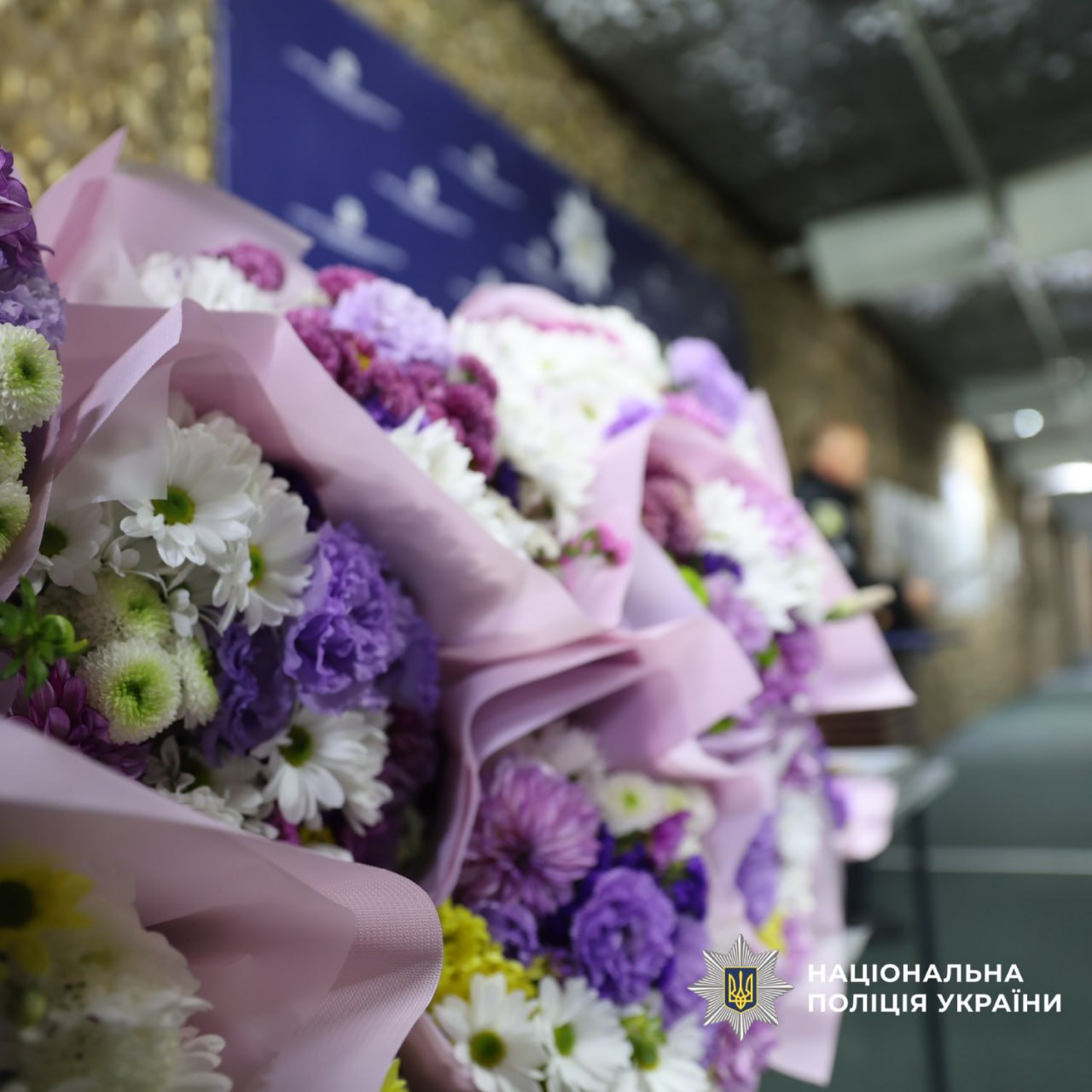 Великий букет квітів, загорнутий у рожевий папір, з фіолетовими, білими та жовтими хризантемами.