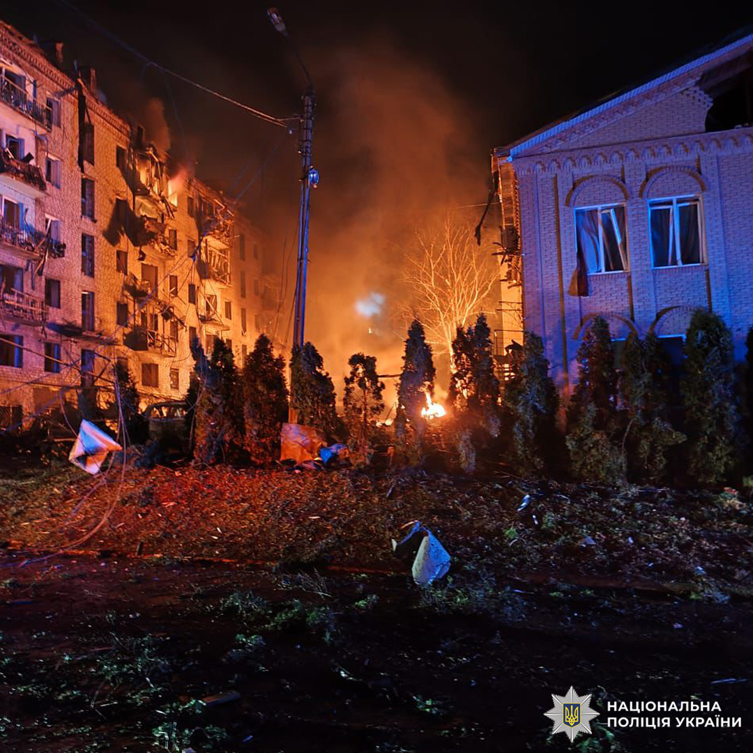 Пожежа після обстрілу в житловому районі: одна будівля охоплена вогнем і сильно пошкоджена, інша — освітлена аварійними вогнями; між ними — дерева, уламки та сліди вибуху.