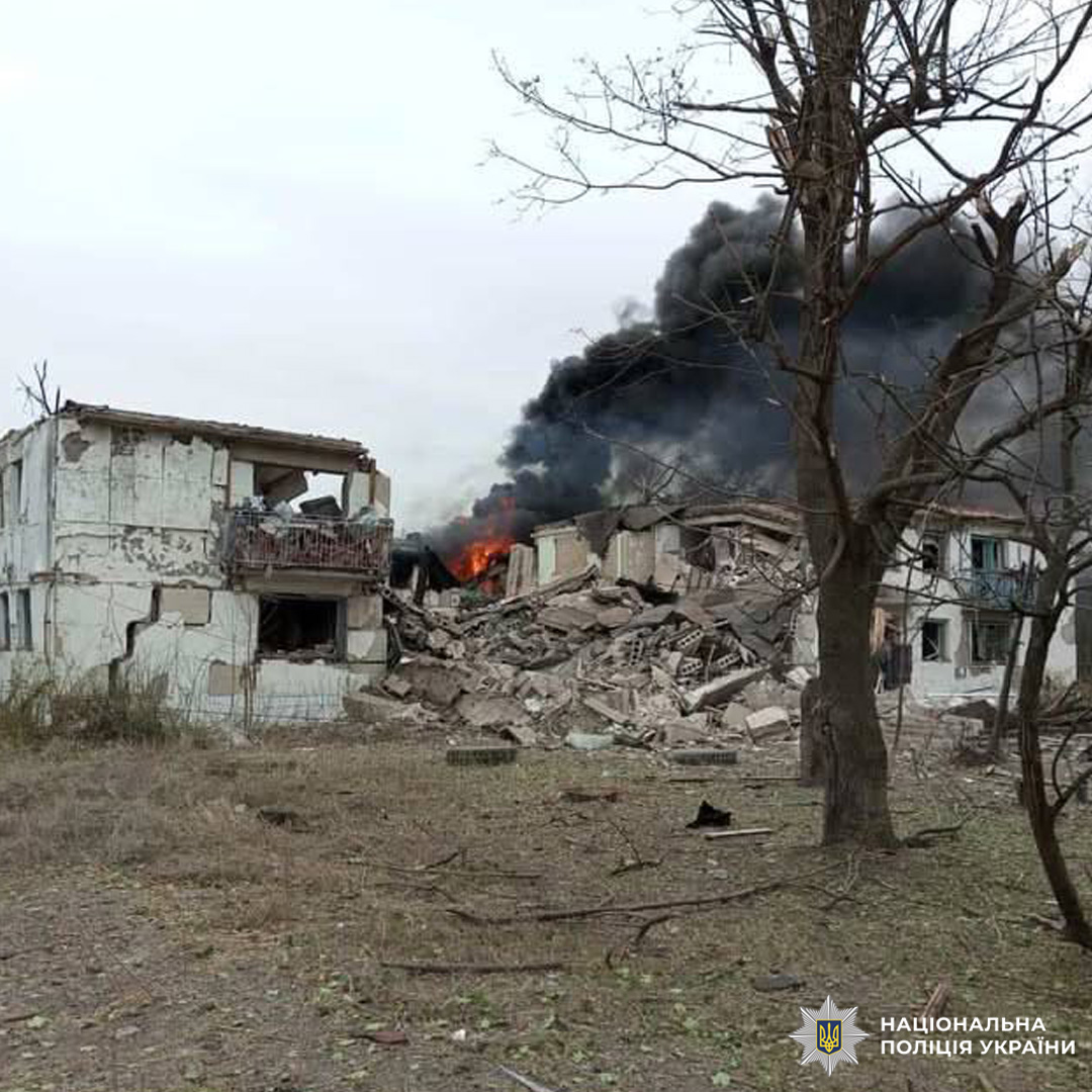 Зруйновані будівлі. Одна частково обвалена, поруч — велика пожежа з густим чорним димом. На передньому плані — суха трава, голі дерева.