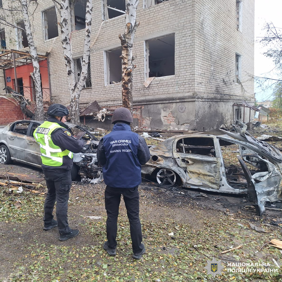 Поліцейський і прокурор з написом "WAR CRIMES PROSECUTOR" оглядають згоріле авто біля пошкодженої багатоповерхівки, навколо уламки.
