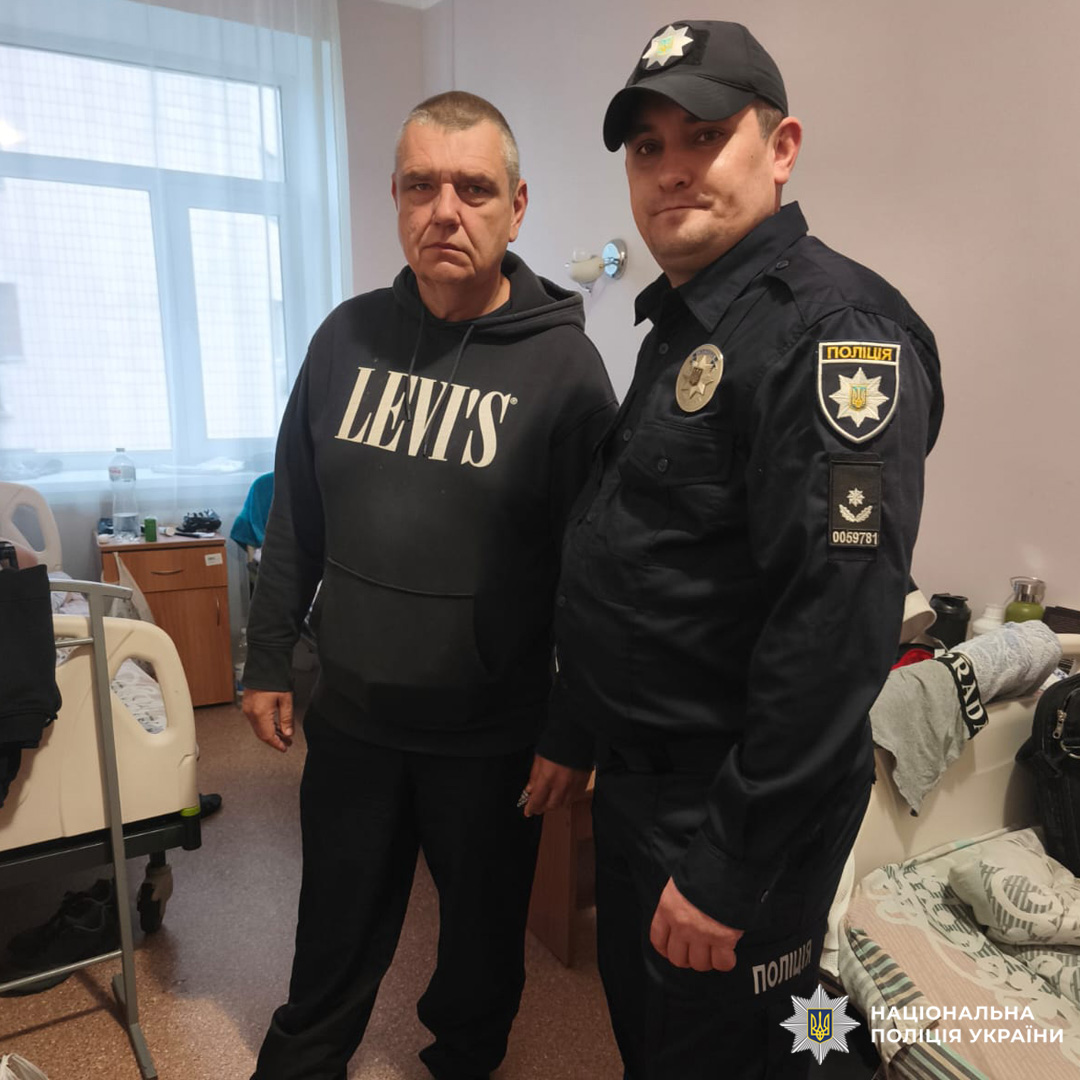 Двоє людей у лікарняній палаті. Один у худі з написом "LEVI'S", другий у формі поліцейського. На фоні — два ліжка, стіл з речами та вікно з прозорими шторами.