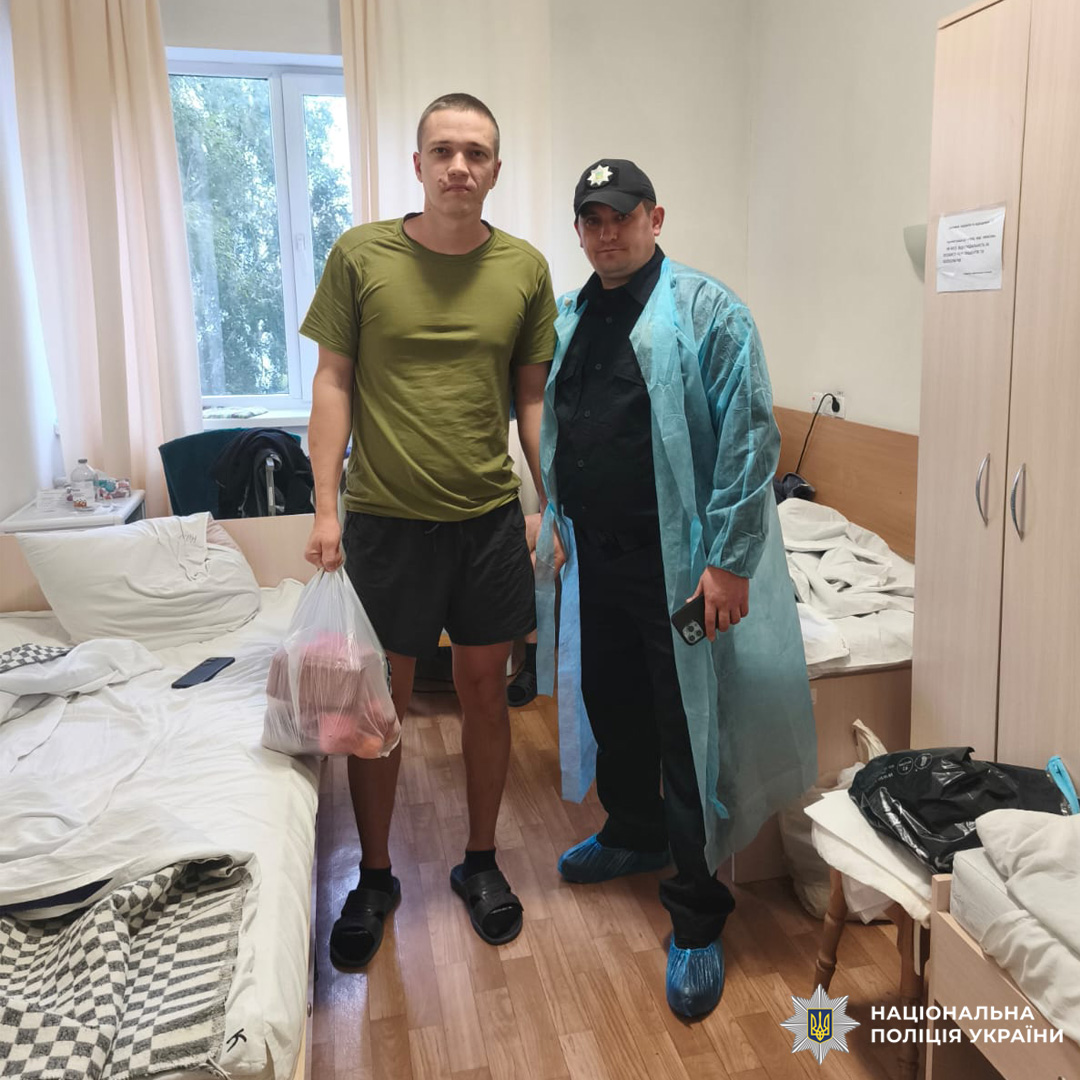 Двоє у палаті. Один у зеленій футболці тримає пакет, інший — у чорному одязі з синім медичним халатом і бахілами; на фоні — два ліжка з білою білизною, дерев’яна підлога, меблі та вікно з білими шторами.
