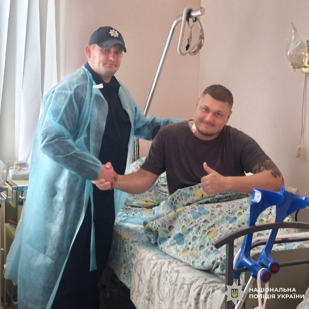 Поранений боєць у лікарняному ліжку тисне руку поліцейському в формі з медичним халатом. На руці татуювання, поруч медичне обладнання.