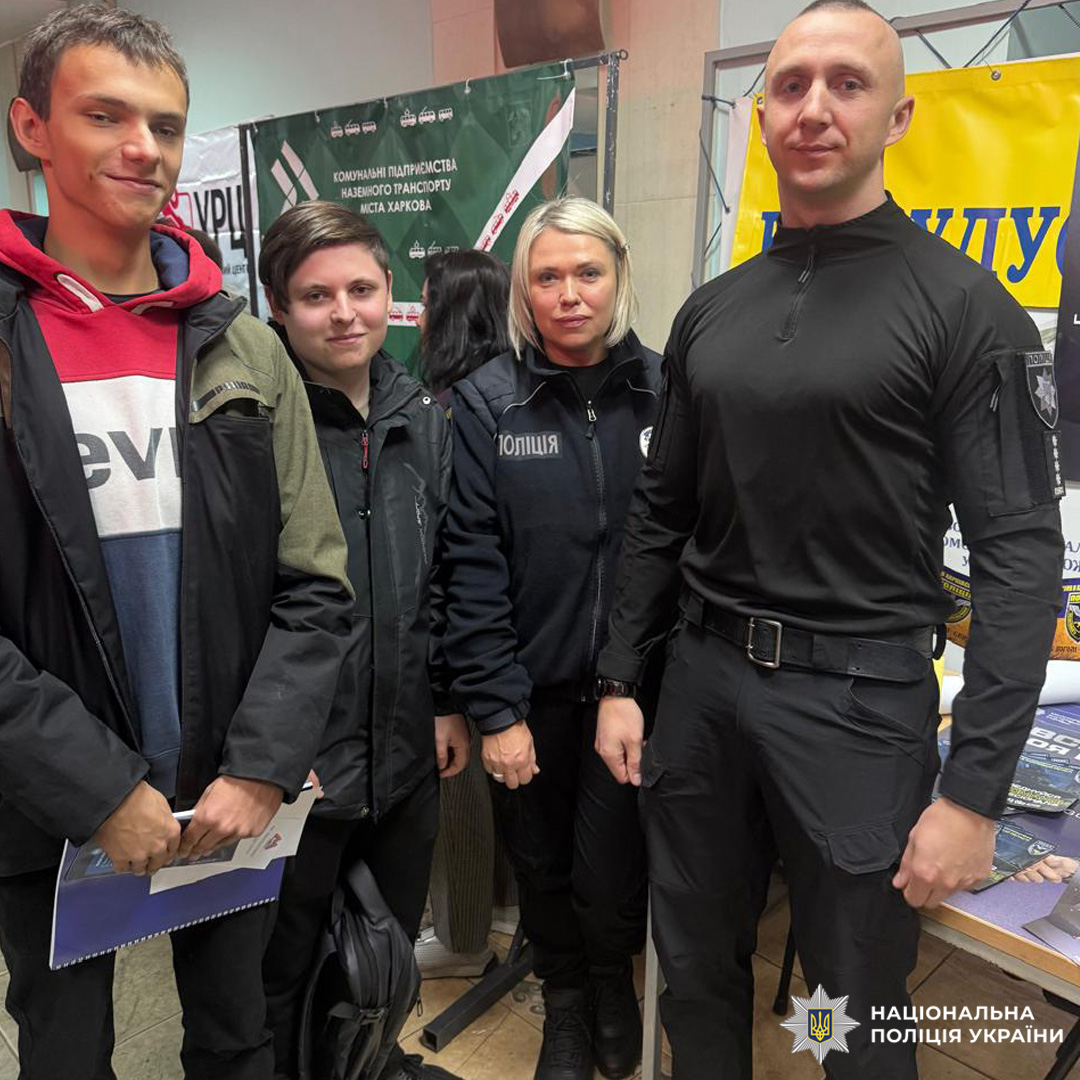 Люди на тлі інформаційних стендів: двоє — у формі Поліція, троє — у повсякденному одязі; сцена схожа на освітній або профорієнтаційний захід.