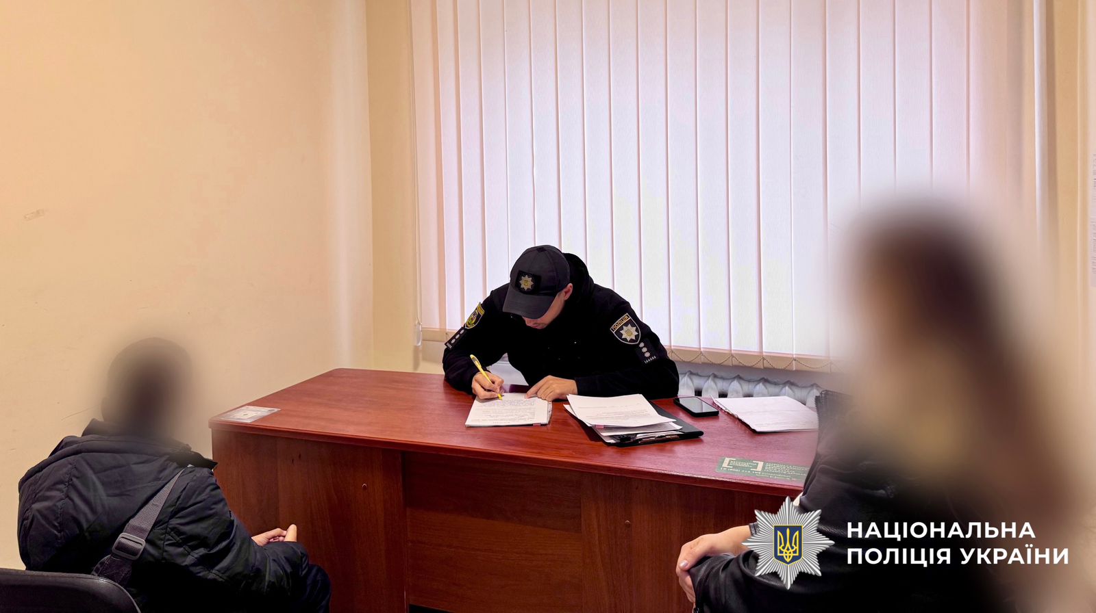 На фото зображений поліцейський, який складає адміністративні матеріали. Навпроти нього за столом сидять малолітній та його матір.