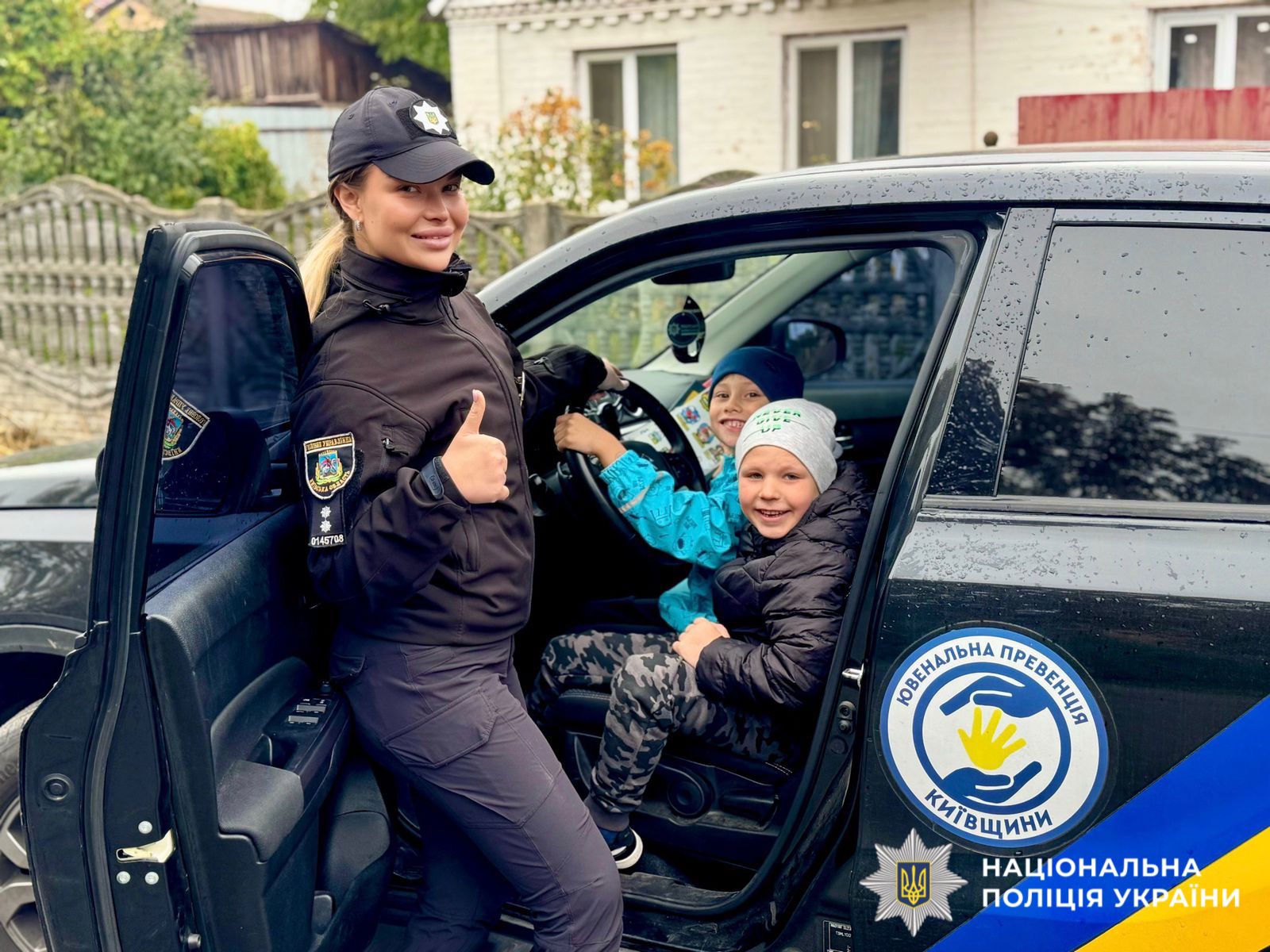 На фото зображені діти у поліцейькому автомобілі на сидінні водія. Двері авто відкриті. Поряд, біля автомобіля стоїть поліцейська.