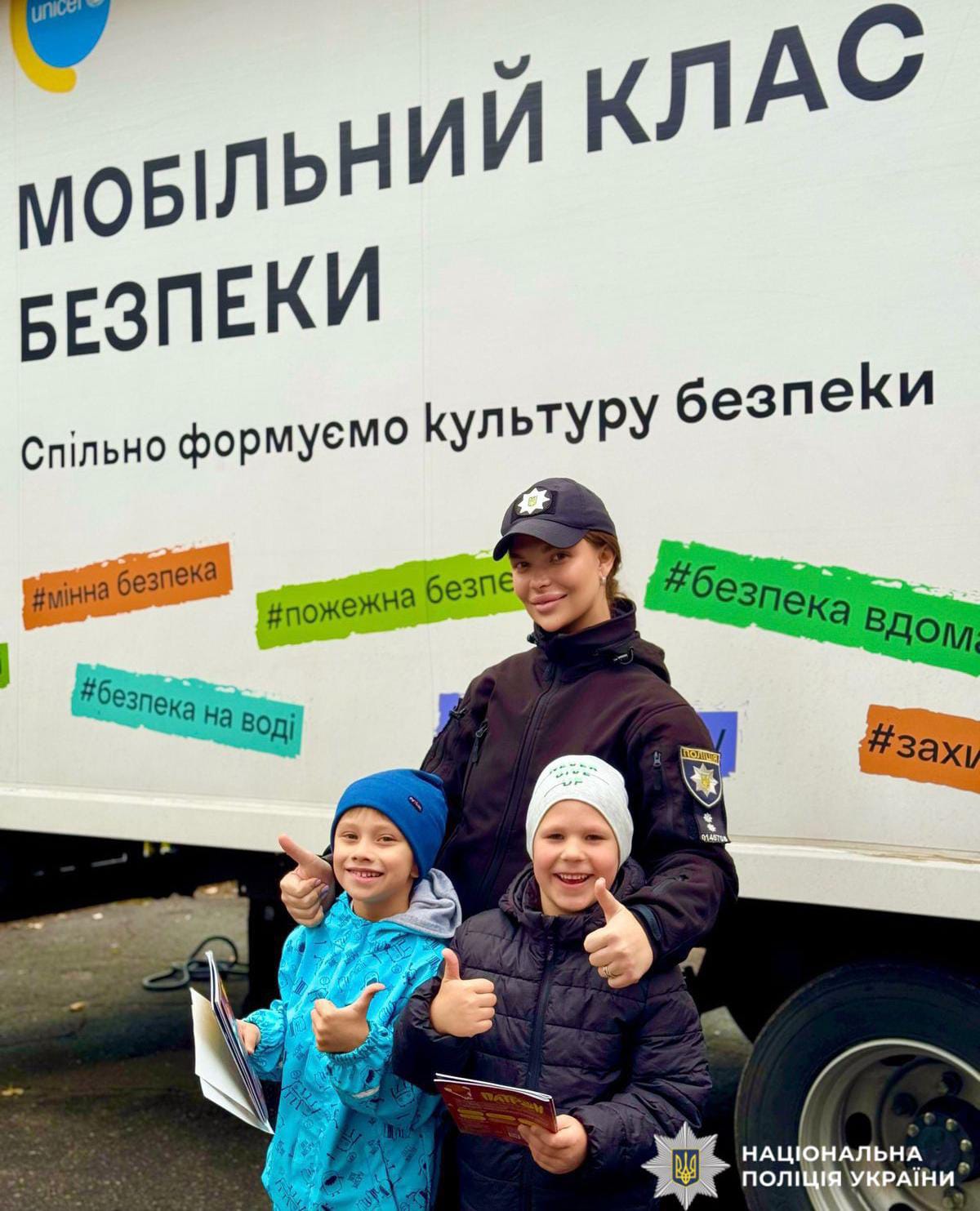 Поліцейська стоїть за дітьми. Всі троє на фоні мобільного класу безпеки.